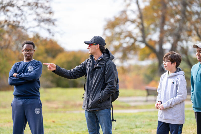 The image shows a group of people in a park. A man in a black jacket is pointing to his left. There are three other people in the frame, one of them is looking at the man in the black jacket. The background shows trees and a bench.